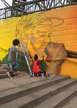 Youth apprentice Alex Baez Rivera looks on as artist Brittany Williams works on a section of the mural featuring Moses Rockwell. - PHOTO BY REBECCA RAFFERTY