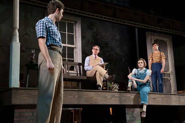 An older man and a younger man and younger woman up on the stage look down on a young man standing in the foreground. The stage is dark with spotlights illuminating the characters.
