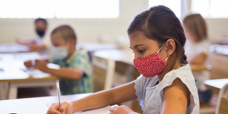 A young student wears a cloth face mask at their school desk.