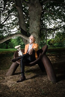 Wendell Castle, photographed outside his Scottsville studio in 2016, seated on a chair of his own creation and holding a scale model of another sculpture.